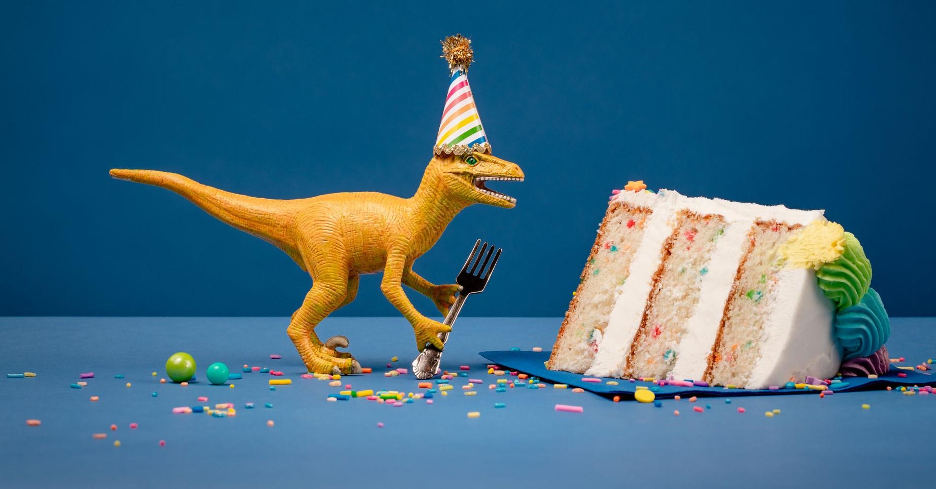 Toy Dinosaur holding a fork next to a slice of birthday confection on a undecorous background.